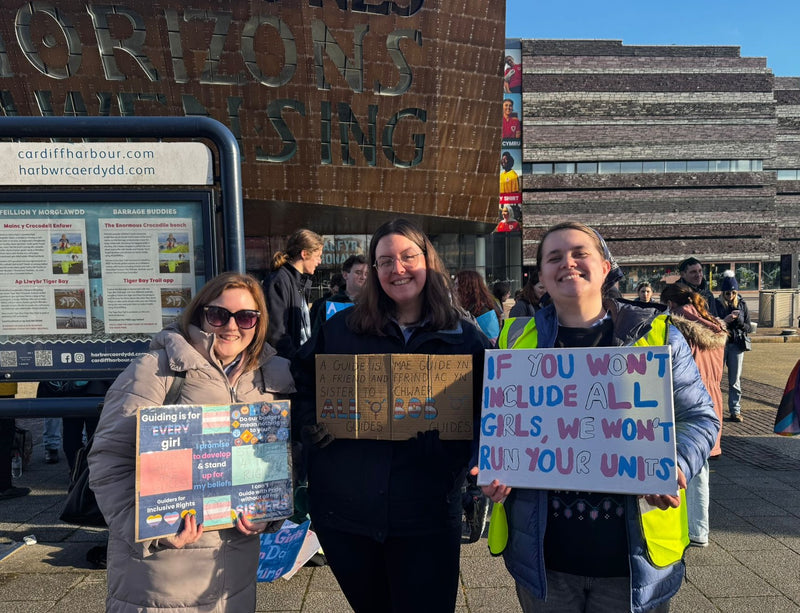 Three protesters outside Wales Millennium Centre holding up signs, one of which is in English and Welsh