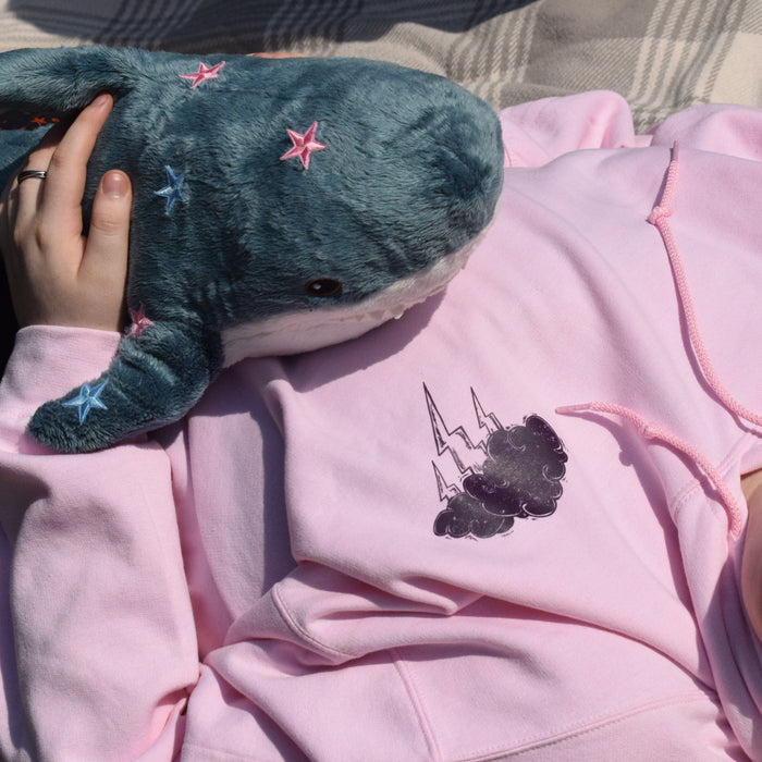 A person lying on a plaid blanket outdoors, wearing a light pink hoodie with a dark graphic thunder cloud print on the left chest and holding a blue plush whale toy with small pink and blue star details.