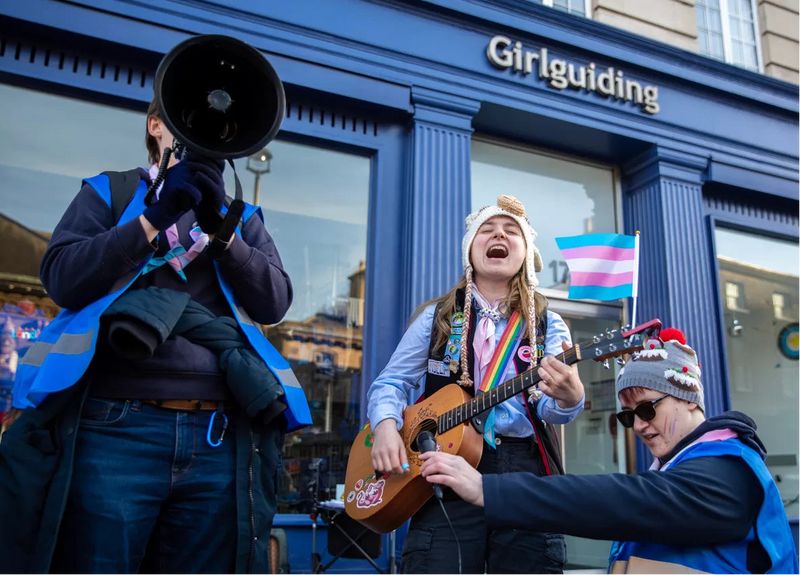 Three protesters in front of Girlguiding HQ with a megaphone and guitar