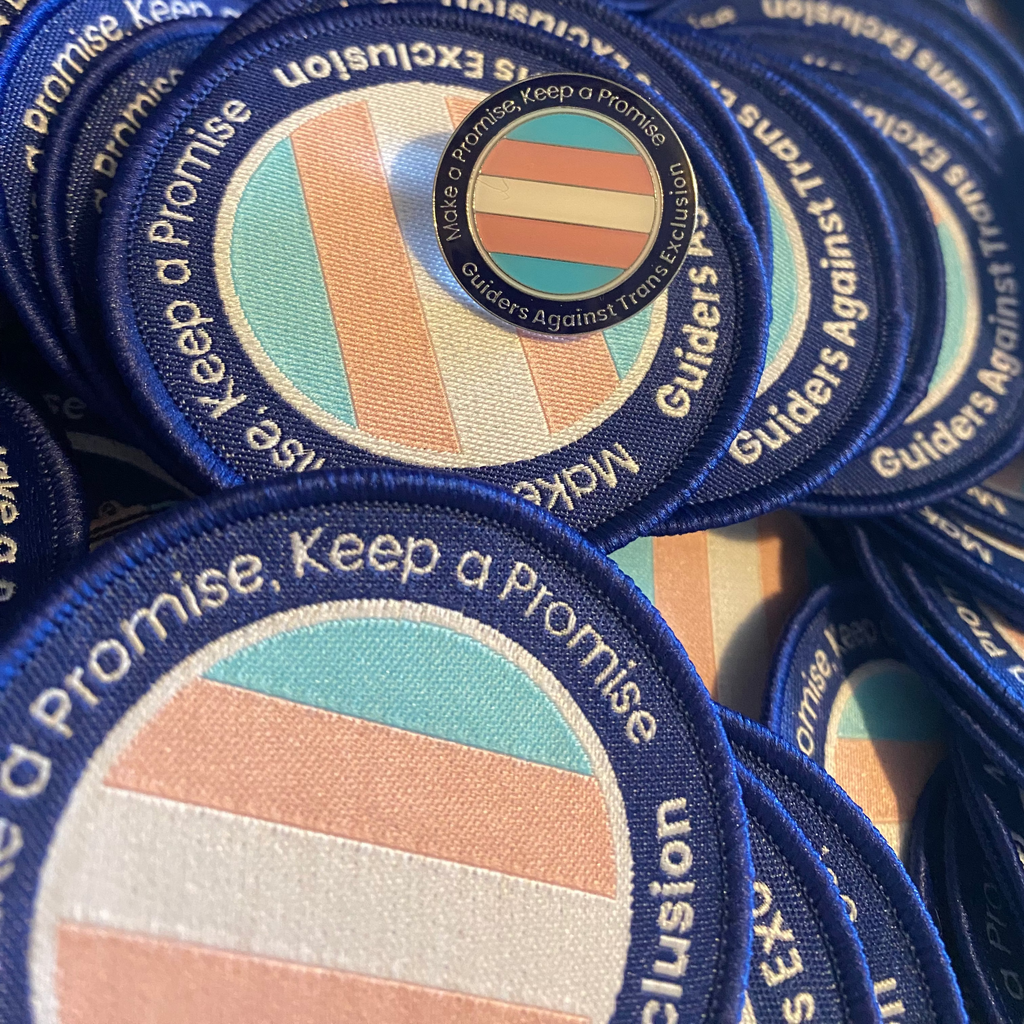 A pile of guiders against trans exclusion logo woven badges, with an enamel pin of the same design on top