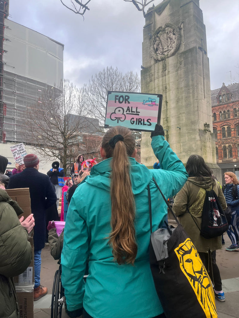 Protester amongst the crowd with their back to the camera holding up a sign with "for all the girls" on a trans flag with our mascot shark, Reef, in the corner