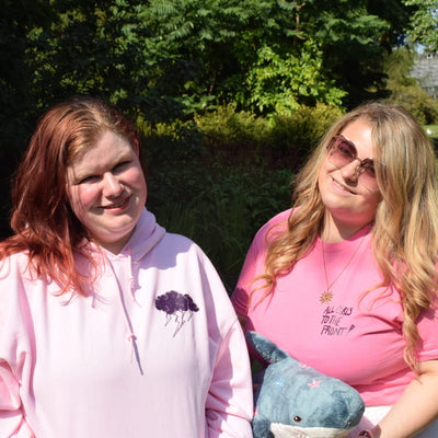 Two women standing outdoors in a sunny park setting. The woman on the left wears a light pink hoodie with thunder cloud graphic on the right, and the woman on the right wears a pink graphic t-shirt saying " all girls to the front" while holding a blue plush shark toy decorated with white and pink stars .