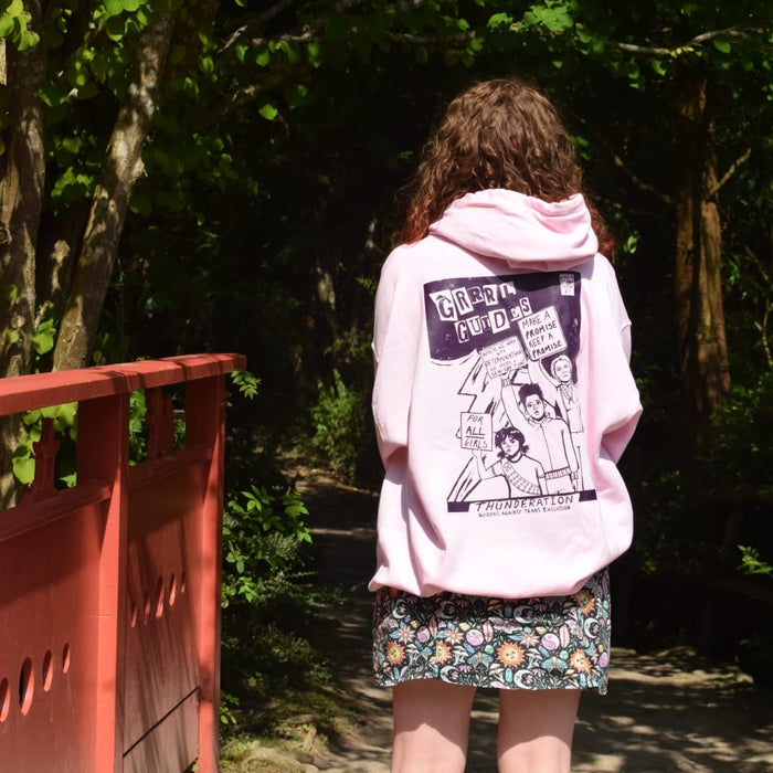 A person standing outdoors on a path in front of a red bridge, viewed from behind. The person is wearing a light pink hoodie with a large protest inspired graphic print on the back and a patterned skirt.