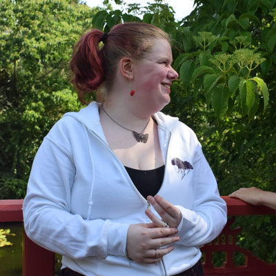 A woman with red hair in a ponytail, wearing a white zip-up hoodie over a black top and a dark patterned skirt, standing outdoors on a red railing with green foliage in the background.