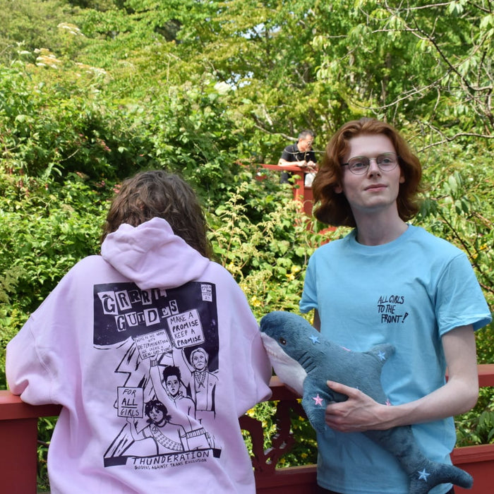 Two people standing outdoors on a red railing in front of dense green foliage. The person on the left wears a light pink hoodie with a large protest graphic print on the back and the person on the right wears a vibrant sky blue t shirt with  " All girls to the front written on the upper left of the chest.