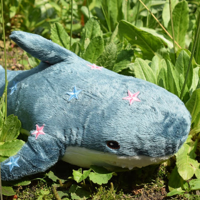 A plush shark toy in blue-gray fabric with embroidered pink and blue stars and a white underbelly, photographed outdoors among green plants.
