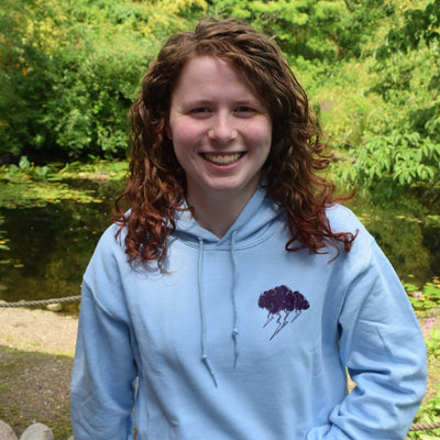 A smiling young woman with curly brown hair stands outdoors in front of a garden pond, wearing a light blue hoodie with a small dark purple cloud graphic on the left chest and a floral skirt.